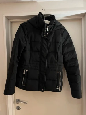 Svart quiltad pufferjacka med huva - Snygg svart pufferjacka med quiltad design och avtagbar huva. Jackan har hög krage, dragkedja framtill och två fickor med dragkedjor. Perfekt för kalla dagar och ger en modern look.