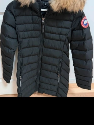 Svart dunjacka  med pälskrage - Snygg svart dunjacka  med luva och pälskant. Jackan har två dragkedjeförsedda fickor fram, quiltad design och klassisk logopatch på ärmen. Perfekt för kalla vinterdagar och riktigt varm tack vare dunfyllningen. Funkat på Xs, S äkta päls, helt ny