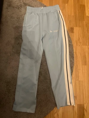 Palm Angels ljusblå mjukisbyxor - Säljer ett par ljusblå mjukisbyxor från Palm Angels med vita och svarta sidoränder. Byxorna har resår i midjan, raka ben och dragkedja vid benslut. Snygg logga på vänster sida och två fickor fram. Perfekta för en chill och trendig look. QR finns på byxorna