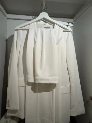 Vit kostym med kavaj och byxor - Säljer en stilren vit kostym med klassisk kavaj och matchande byxor. Kavajen har enkel knappstängning, två fickor framtill och smala slag. Perfekt för dig som vill ha en clean och modern look. Byxorna är vida! Kavajen är oversize i storlek S och byxorna vida i storlek 34!