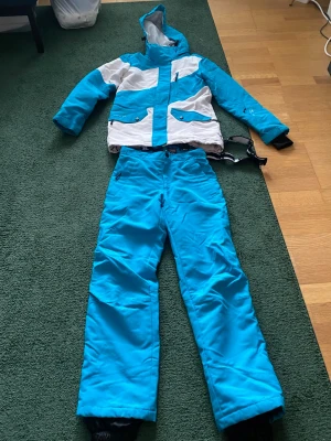 Blått skidset med jacka och byxor - Säljer ett blått skidset från Everest med matchande jacka och byxor, använd 1 gång.Jackan har vit och blå färgblockdesign, huva och flera fickor med dragkedja. Byxorna är helblå med justerbar midja och dragkedja. Perfekt för vinterns skidäventyr.
