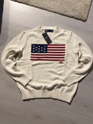 Vit stickad tröja Ralph Lauren - Stickad vit tröja från Ralph Lauren med amerikansk flagga i blått, rött och vitt på bröstet samt RL-broderi. Tröjan har rund halsringning och ribbade muddar vid ärmslut och nederkant. Perfekt för dig som gillar klassisk och preppy stil.