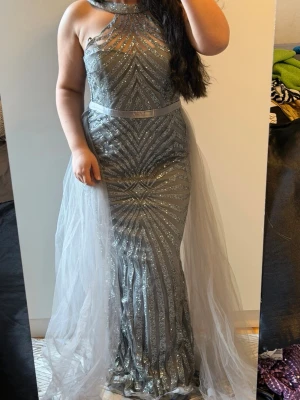Glittrig silver exklusiv långklänning - Säljer en glittrig silverfärgad långklänning med geometriskt mönster och transparent mesh upptill. Klänningen har en markerad midja med satinband och ett lager av tunn tyll som ger extra volym. Perfekt för dig som vill sticka ut på bal eller fest.