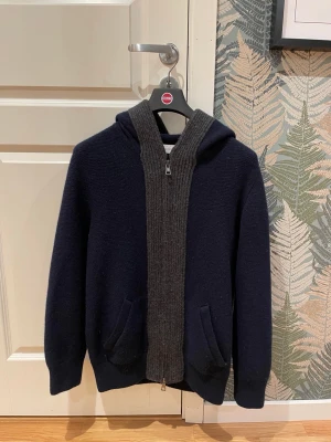 Marinblå cardigan/hoodie från Moncler - Snygg marinblå hoodie från Moncler med grå stickad dragkedja och huva. Tröjan har två fickor framtill och en Moncler-logga på armen.