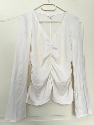 Vit långärmad blus med rynk framtill - Trendig vit blus med djup V-ringning och rynkdetalj längs magen. Långa ärmar och lätt utsvängd nederkant ger en snygg siluett. Tillverkad i ett glansigt, satinliknande material som känns lyxigt och passar till många olika stilar. Har märken uppe vid höger axel, går evetuellt bort i tvätt. 