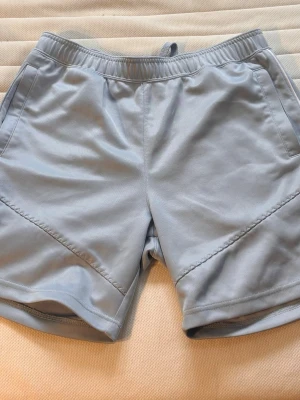 Ljusblå träningsshorts från Nike - Snygga ljusblå träningsshorts från Nike med elastisk midja och snörning. Shortsen har två sidofickor och är tillverkade i ett mjukt och lätt syntetmaterial som passar perfekt för aktiva dagar.