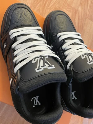 Louis Vuitton svarta sneakers - Snygga svarta Louis Vuitton sneakers med vita detaljer och vita skosnören. Skorna har LV-logga på plösen, sidan och hälen samt präglade monogramdetaljer. Tillverkade i läder och textil med robust sula och cool design.