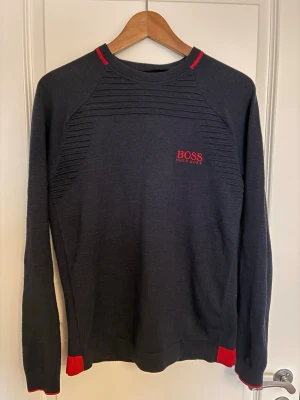Marinblå tröja från Hugo Boss - Snygg marinblå tröja från Hugo Boss med röda detaljer vid hals, ärmslut och nederkant. Tröjan har diskreta ribbade partier upptill och röd logotyp på bröstet. Perfekt för en stilren och modern look.