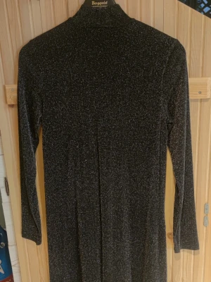 Svart glittrig långärmad klänning - Snygg svart klänning med glittereffekt och lång ärm. Klänningen har en rak passform och hög hals, perfekt för dig som vill sticka ut lite extra. Materialet känns mjukt och stretchigt, vilket gör den bekväm att bära.