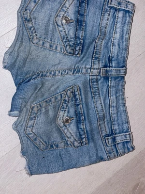 Blå jeansshorts med rå kant - Säljer ett par blå jeansshorts med råa kanter och slitningar. Shortsen har stora fickor med knappdetaljer bak och klassisk femficksdesign fram. Perfekta för sommaren och ger en avslappnad streetstyle-look.
