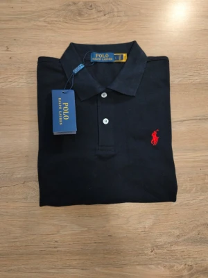 Svart pikétröja från Polo Ralph Lauren - Klassisk svart pikétröja från Polo Ralph Lauren med röd broderad logga på bröstet. Tröjan har krage, knappar framtill och korta ärmar. Tillverkad i mjuk bomull som känns skön mot huden. Perfekt för en stilren och avslappnad look. Det står att den är Large men den är i storleken Small 