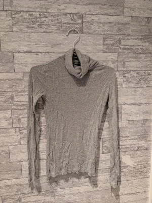 Grå ribbad polotröja - Säljer en tunn och ribbad grå polotröja med lång ärm. Tröjan har en hög krage och är figurnära i modellen, perfekt att styla med jeans eller kjol. Materialet är mjukt och stretchigt, vilket gör den bekväm att bära.