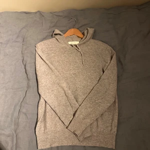Grå cashmere hoodie från Sayless - Mjuk och lyxig grå hoodie från Sayless i cashmere. Tröjan har huva med snörning och långa ärmar, perfekt för dig som gillar chill och stilren look. Ribbstickade muddar vid ärmslut och nederkant ger extra komfort. Designad i Sverige. Passar 170-175