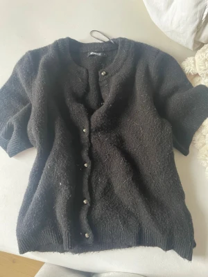 Svart kofta från Gina Tricot - Kortärmad svart kofta från Gina Tricot med rund halsringning och dekorativa silverfärgade knappar framtill. Koftan är stickad i ett mjukt material och har ribbade muddar vid ärmslut och nederkant. Perfekt för lager på lager och enkel att styla.