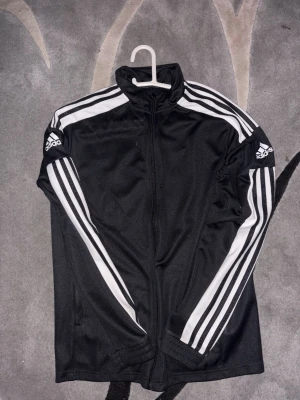 Svart Adidas träningsjacka - Säljer en svart träningsjacka från Adidas med vita klassiska ränder längs ärmarna och Adidas-logga på båda axlarna. Jackan har dragkedja framtill, hög krage och är tillverkad i ett glansigt syntetmaterial. Perfekt för träning eller chill.