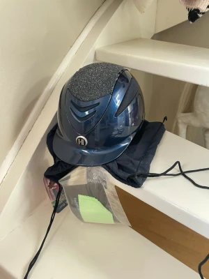 Mörkblå One K ridhjälm med glitter - Ordinarie pris runt 3500kr. Säljer en snygg mörkblå ridhjälm från One K med glittrig topp och ventilerande detaljer. Skal strl 0 alltså så passar en liner 47-54. Aldrig använd och helt ny med lapp, har ingen liner som ingår men har en utbytbar topp-panel som ingår så man kan byta(se sista bilden). Hjälmen har en modern design och kommer med tillhörande förvaringspåse. Perfekt för dig som vill sticka ut i stallet! Skriv om du har några frågor! 