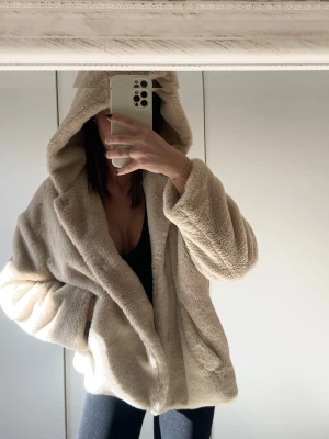 Beige teddyjacka med huva - Supermjuk och fluffig teddyjacka i beige med stor huva och avslappnad passform. Jackan har långa ärmar och två sidofickor, perfekt för att hålla sig varm och snygg under kyliga dagar. Materialet är fuskpäls som ger en mysig känsla.