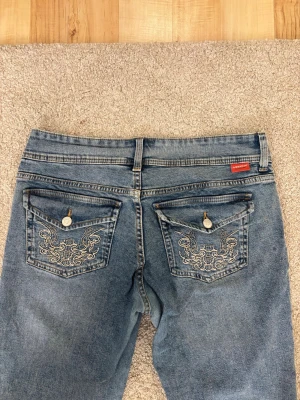 Blåa jeans med broderade fickor - Säljer ett par blåa jeans från H&M med snygga broderier på bakfickorna och dekorativa knappar. Jeansen har klassisk passform och är gjorda i denim. Perfekt för dig som gillar detaljer och vill sticka ut lite extra. Lite tunna i grenen men knappt använt. 
