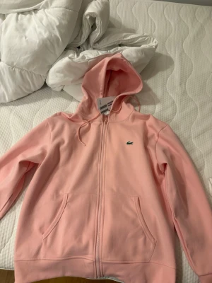 Ljusrosa hoodie från Lacoste - Snygg ljusrosa hoodie med dragkedja från Lacoste. Klassisk krokodillogga på bröstet, två fickor framtill och huva med dragsko. Mjuk och skön bomullsblandning, perfekt för en chill och avslappnad stil.