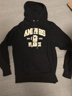 Svart hoodie från Ami Paris - Assnygg svart hoodie från AMI PARIS. Tröjan är i storlek M och är bara använd ett fåtal gånger. Säljer då jag tömmer min garderob på kläder jag sällan använder. Skriv vid vilka som helst funderingar, pris kan diskuteras!