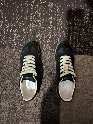 Maison margiela gats - Snygga svarta sneakers med vita snören och beige sula. Klassisk låg modell med stilrena linjer och kontrasterande detaljer. Ovandelen är i skinn och mocka för en cool och tidlös look. Perfekta för dig som gillar minimalistisk stil.