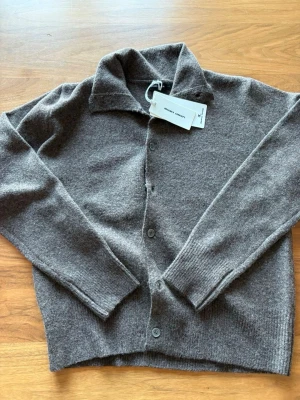 Brun cardigan Decency Concept - Mysig brun cardigan från Decency Concept i mjuk ullmix. Klassisk design med knappar framtill, ribbade muddar och krage. Perfekt för lager på lager och enkel att matcha med olika outfits.