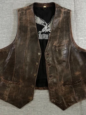 Vintage leather biker vest in heavy genuine leather 🤎 - Deep brown distressed patina with real character – not fake factory distress.  Classic 5-button front, chest pocket + adjustable back strap. Perfect layered over hoodie or tee. Proper Americana / biker vibe.  Inner lining hem slightly coming loose in one area (see photo). Does not affect wear.  Measurements (flat): S2S 35 cm P2P 57 cm Front length 59.5 cm Waist 55 cm  Good vintage condition with natural wear throughout. Item #75