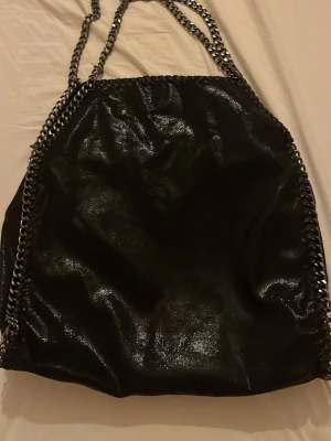 Svart kedjeväska från Stella McCartney - Cool svart handväska i glansigt veganskt läder med silverfärgade kedjedetaljer längs kanterna och som axelrem. Väskan har en mjuk, slouchy form och är perfekt för dig som gillar statement-accessoarer. Helt oandvänd.