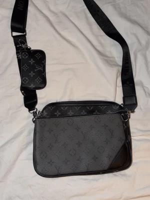 Louis Vuitton svart axelväska monogram - Snygg svart axelväska från Louis Vuitton med klassiskt monogrammönster. Väskan har en bred justerbar axelrem och en mindre avtagbar ficka framtill. Tillverkad i läder med silverfärgade detaljer och dragkedja. Perfekt för dig som vill ha en trendig och praktisk väska.