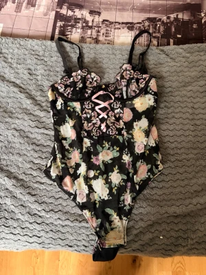 Blommig spetsbody med korsettband - Snygg body i svart mesh med blommigt mönster och detaljer i rosa spets. Framtill finns dekorativa korsettband i ljusrosa och kuporna har broderade blommor. Justerbara axelband och genomskinligt material ger en feminin och trendig look. Helt ny med prislapp