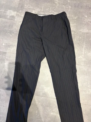 Svarta kostymbyxor med kritstreck - Snygga svarta kostymbyxor med tunna vita kritstreck. Klassisk rak passform med hällor för bälte, knapp och dragkedja fram samt två bakfickor med knapp. Perfekta för en stilren och dressad look.