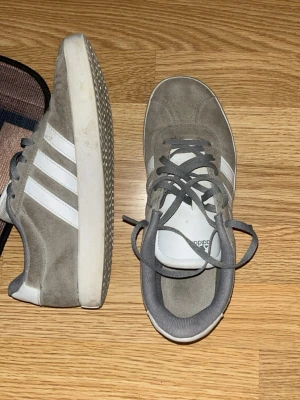 Grå Adidas sneakers i mocka - Snygga grå sneakers från Adidas i mocka med klassiska vita ränder på sidorna och vit sula. Lågt skaft och gråa skosnören. Perfekta för dig som gillar stilrena och sportiga skor med retrovibe.