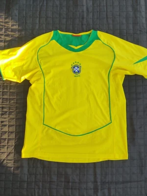 Gul Brasilien fotbollströja använd bara för VM 2006 - Snygg gul fotbollströja med gröna detaljer och Brasilien-emblem på bröstet. Tröjan har korta ärmar, grönt parti vid halsen och är tillverkad i ett lätt och ventilerande material, perfekt för träning eller för att gå och se på match där Brasilien spelar. Använd enbart under VM 2006.