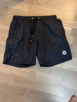 Svarta shorts från Moncler med snörning - Säljer ett par svarta shorts från Moncler med dragsko i midjan och Moncler-logga på vänster ben. Shortsen har en enkel design och är perfekta för sommaren. Materialet känns lätt och syntetiskt.