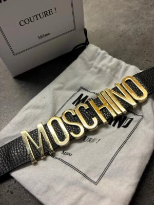Svart skinnarmand  med gulddetaljer från Moschino - Skavanker på guld texten , därav priset . Forskande mycket att ge . Äkta köpt på raglady . Finns ej kvitto men på boxen är prislappen som man ser tydligt . Tillkommer dustbag med .