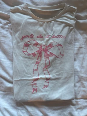 Vit t-shirt med rosa rosetttryck - Vit t-shirt med rund hals och tryck i rosa framtill med texten 'girls do it better' och en stor rosett.