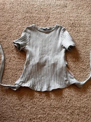 Ljusgrå ribbad topp med knytband - Trendig ljusblå ribbad topp med korta ärmar och snygga knytband bak som ger en unik look. Toppen har rund halsringning och är tillverkad i mjukt bomullsmaterial. Perfekt för dig som gillar stilrena detaljer och en modern vibe.