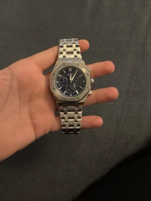 Silverfärgad kronografklocka i stål - Stilren klocka med silverfärgat armband i rostfritt stål och mörkblå urtavla med flera små urtavlor. Klockan har en sexkantig boett med synliga skruvar och kronograf-funktion. Perfekt accessoar för dig som gillar en sportig men ändå elegant look.