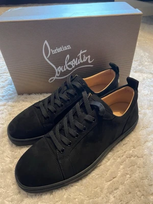 Svarta sneakers från Christian Louboutin - Stilrena svarta sneakers från Christian Louboutin i mocka med klassisk röd sula och svarta snören. Snygg och minimalistisk design med diskreta detaljer och lyxig känsla. Perfekta för dig som vill sticka ut med exklusiva skor.