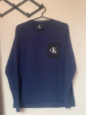 Mörkblå långärmad topp från Calvin Klein - Mörkblå långärmad topp från Calvin Klein med rund halsringning och svart bröstficka med vit CK-logga. Enkel och stilren design som passar till många olika outfits. Tillverkad i mjukt material för en bekväm känsla.