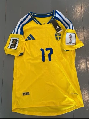 Sverige matchtröja Gyökeres #17 Adidas - Gul och blå officiell Sverige matchtröja från Adidas med Gyökeres och nummer 17 på ryggen. Tröjan har FIFA och VM-märken på ärmarna, klassiska Adidas-ränder på axlarna och Climalite-material som andas. Perfekt för fotbollsfans som vill sticka ut på läktaren.