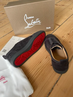 Christian Louboutin gråa sneakers - Snygga gråa sneakers från Christian Louboutin i mocka med klassisk röd sula och diskreta detaljer. Skorna har lågt skaft, svarta skosnören och exklusiv känsla. Perfekta för dig som vill sticka ut med en ikonisk design och lyxig touch. HELT OANVÄNDA HELT NYA 10/10 skick loubs. Alla tillbehör är medföljer. Pris kan diskuteras 