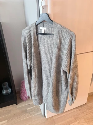 Grå ribbstickad kofta från H&M - Mysig grå kofta från H&M i ribbstickat material. Koftan har öppen front utan knappar, långa ärmar och en avslappnad passform. Perfekt att slänga på sig över en t-shirt eller topp för en chill look.