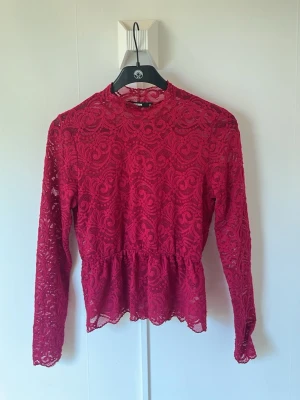 Vinröd spetsblus med lång ärm från BIKBOK - Snygg vinröd blus i spets med lång ärm och hög hals. Blusen har ett genombrutet mönster och volang nedtill som ger en feminin look. Perfekt att styla med jeans eller kjol för en trendig outfit. Jättesnygg på. 
