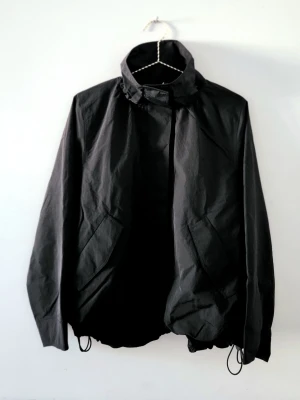 COS Svart nylonjacka med hög krage Utsåld online - Modern jacka som föreslår en subtil tolkning av utilitydesign, sydd i nylon med klassisk, svart nyans. Ledigt snitt med hög, rynkad krage, fickor med lock och resår och dragsko i nederfållen, så att du kan experimentera med silhuetten.   