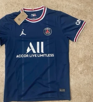 PSG matchtröja blå Jordan x Nike - Snygg blå Paris Saint-Germain matchtröja med vita och röda detaljer på krage och ärmslut. PSG-logga på bröstet, Jordan-logga och sponsortryck framtill. Tillverkad i lätt och ventilerande material, perfekt för fotboll eller träning.