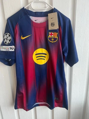 FC Barcelona matchtröja Nike - Säljer en FC Barcelona matchtröja i blått och rött med gula detaljer. Tröjan har klubbmärke, Nike-logga, Spotify-logga och Champions League-märke på ärmen. På ryggen står det 'ROONY' och nummer 19 i gult. Materialet är lätt och andas, perfekt för fotboll.