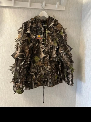 3D camojacka med lövstruktur - Säljer en maskeradjacka med 3D-kamouflagemönster i grönt, brunt och svart. Jackan har lövliknande detaljer som sticker ut för extra realistisk effekt. Storlek S från Fladen 
