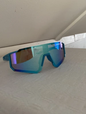 Blå sportiga solglasögon med spegelglas - Snygga blå solglasögon med spegelglas och sportig, kantig design. Bågarna är i blått plastmaterial med coola detaljer och glasen har en reflekterande yta i blå och lila toner. Perfekta för en aktiv livsstil och trendig look.