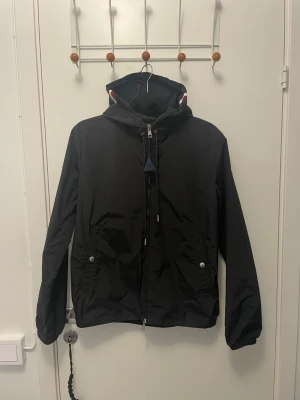 Svart Moncler vindjacka med huva - Snygg svart vindjacka från Moncler med huva och dragkedja framtill. Jackan har Moncler-logga på ärmen och rödvitblå rand på huvan. Två fickor med knappar och justerbar nederkant. Perfekt för blåsiga dagar och streetwear-stil.